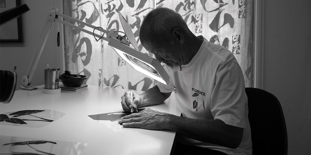 Sig Zane in his studio creating artwork under a lighted magnify glass